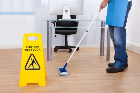 Yellow warning notice to caution people to a slippery wet surface as a janitor mops the floor in an...