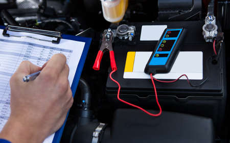 Mechanic's hand writing on clipboard with pliers connected to multimeter on car engine