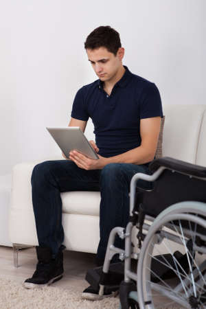 Full length of young handicapped man using digital tablet while sitting on sofa at homeの写真素材