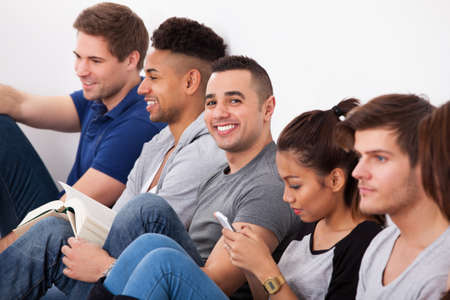 Portrait of happy male college student sitting with classmates against wall in classroomの写真素材
