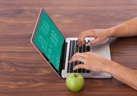 Cropped image of young male student solving math's problem on laptop by green apple on table