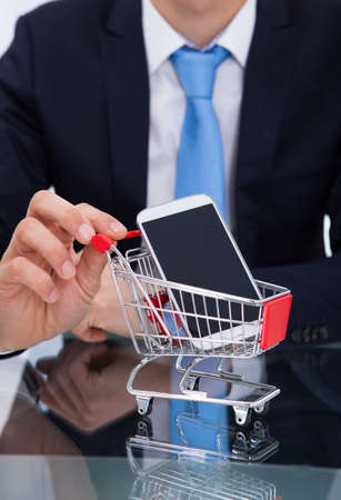 Midsection of businessman pushing smartphone in shopping cart at deskの写真素材