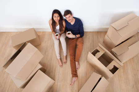 Portrait of young multiethnic couple holding coffee mugs while sitting on floor in new home