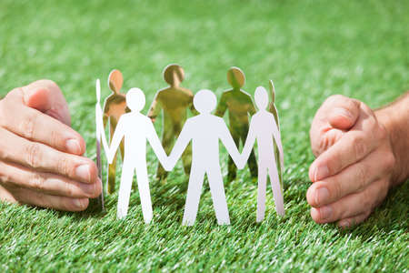 Closeup of hands protecting team of paper people on grassy fieldの写真素材