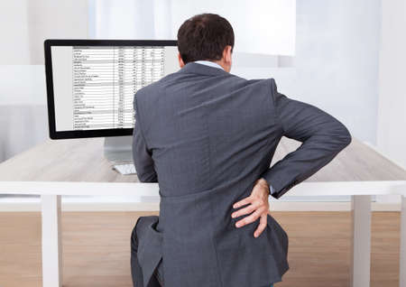 Rear view of businessman suffering from backache while sitting at computer desk in officeの写真素材