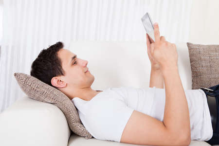 Side view of young man using digital tablet while lying on sofa at home