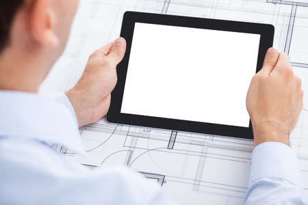 Cropped image of businessman holding blank digital tablet over blueprint in officeの写真素材