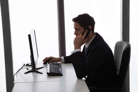 Side view of young businessman answering mobilephone while using computer at desk in officeの写真素材