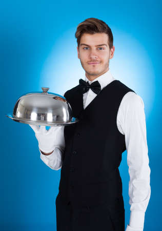 Portrait of confident butler carrying tray over blue background