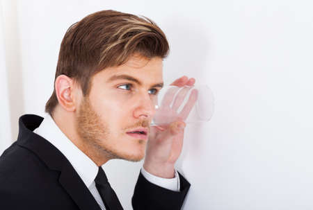 Young businessman with glass on wall eavesdropping in officeの写真素材