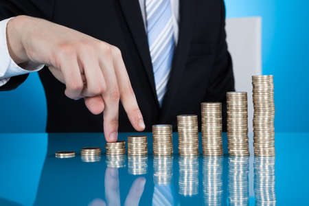 Midsection of businessman's fingers walking up stack of coins at desk against blue backgroundの写真素材