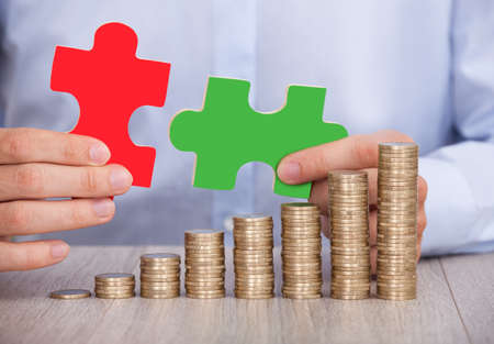 Midsection of businessman with puzzle pieces and stacked coins at office deskの写真素材