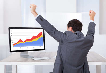Rear view of businessman celebrating success at computer desk in officeの写真素材