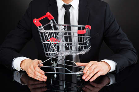 Midsection of young businessman holding shopping cart model at desk against black backgroundの写真素材