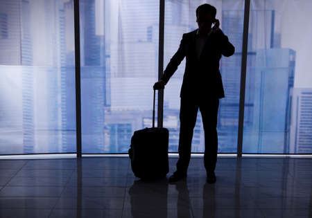 Full length rear view of businessman with luggage using cell phone by office windowの写真素材