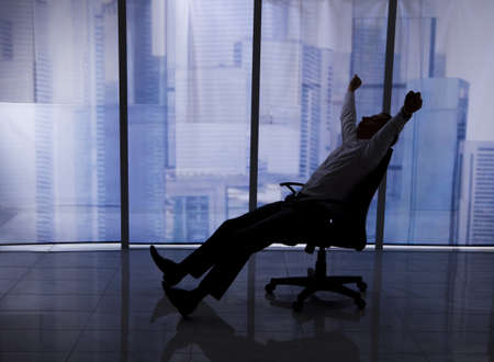Full length of businessman stretching on chair by office window with city view in backgroundの写真素材