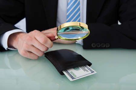 Midsection of young businessman examining wallet with magnifying glass at office deskの写真素材