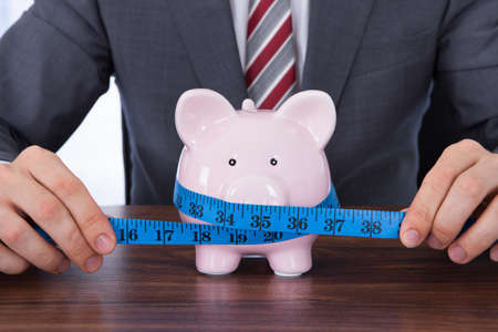 Midsection of businessman measuring piggybank with measure tape at deskの写真素材