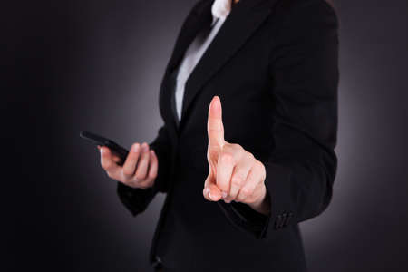 Midsection of young businesswoman pointing while holding cellphone against black backgroundの写真素材