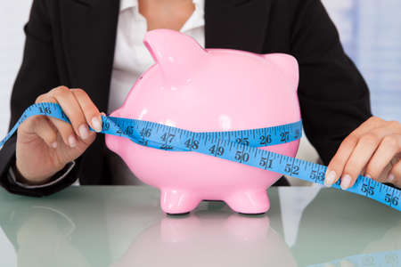 Midsection of businesswoman measuring piggybank with measure tape at deskの写真素材