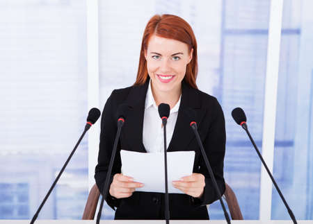 Smiling young businesswoman giving speech on multiple microphones at conferenceの写真素材
