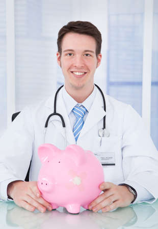 Portrait of young male doctor examining piggybank with stethoscope in hospitalの写真素材