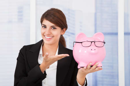 Portrait of happy young businesswoman showing piggybank with eyeglasses in officeの写真素材