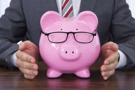 Midsection of businessman shielding piggybank wearing glasses at deskの写真素材