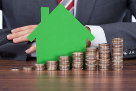 Midsection of businessman with coin stacks by green house model at deskの写真素材