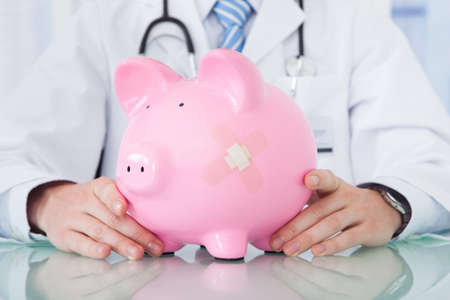 Midsection of young male doctor holding piggybank at desk in clinicの写真素材
