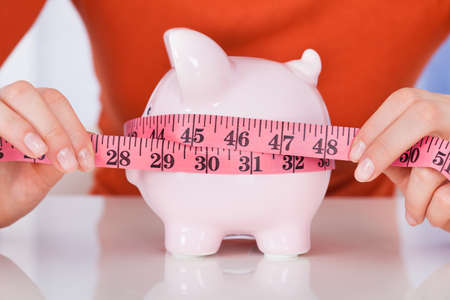 Midsection of young woman measuring piggybank with measure tape at deskの写真素材