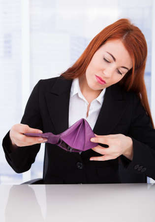 Upset young businesswoman looking at empty wallet while sitting at office desk