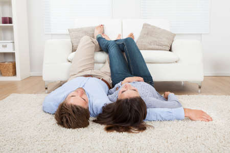 Full length of couple lying on floor with feet up on sofa at homeの写真素材