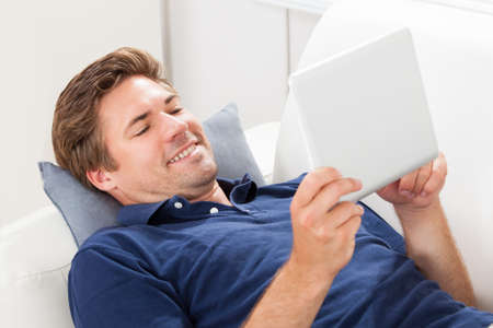 Smiling mid adult man using digital tablet while lying on sofa at home