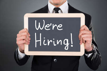 Midsection of young businessman holding slate with We're Hiring sign over gray backgroundの写真素材