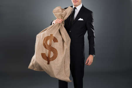 Midsection of businessman carrying sack with dollar sign against gray backgroundの写真素材