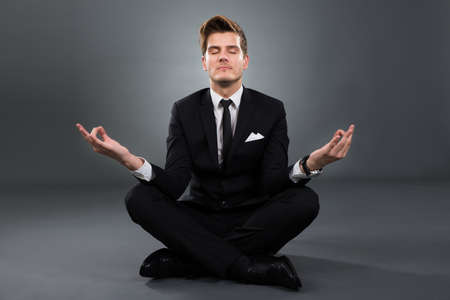 Full length of businessman practicing yoga in lotus position over gray backgroundの写真素材