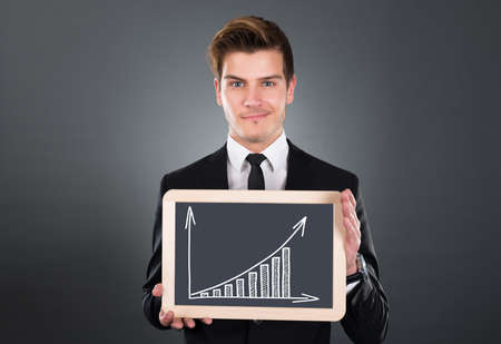 Portrait of young businessman holding slate with growing bar graph over gray backgroundの写真素材