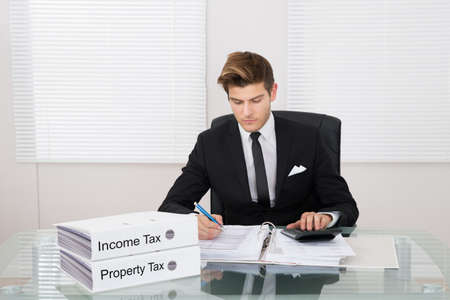 Young businessman calculating tax with binders at desk in officeの写真素材