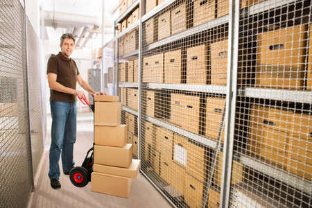 Portrait of delivery man with stacked cardboard boxes on hand truck at warehouse
