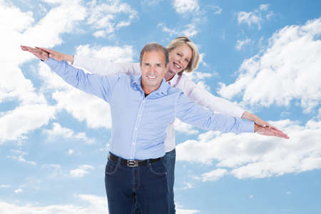 Portrait of happy couple with arms outstretched standing against cloudy sky