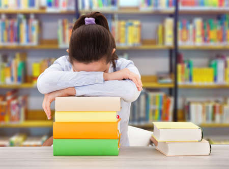 Tired female college student leaning on stack of books at desk in library