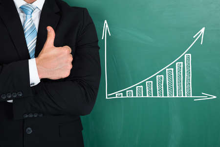 Midsection of young businessman showing thumbs up while standing by graph drawn on chalkboard
