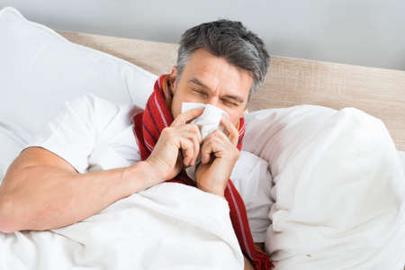 Sick Mature Man Blowing His Nose While Lying On Bed At Homeの写真素材