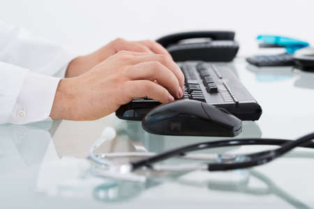 Close-up Of Stethoscope On Desk Besides Doctor Typing On Keyboardの写真素材