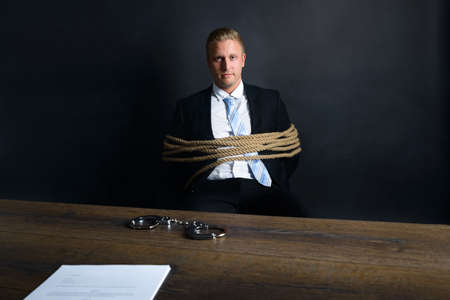 Portrait Of Young Businessman Tied With Rope Sitting In Front Of Table With Handcuff Placed On Itの写真素材