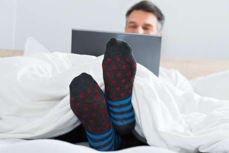 Mature Man With Socks In His Leg Sitting On Bed Using Laptopの写真素材