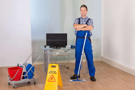 Portrait Of Happy Confident Male Janitor With Cleaning Equipments And Wet Floor Sign In Office
