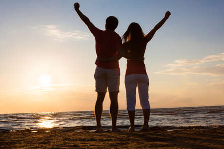Full length rear view of couple with hands raised at beach during sunsetの写真素材
