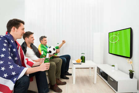 Group Of Friends From Different Nations Holding Beer Bottle Watching Football On Television